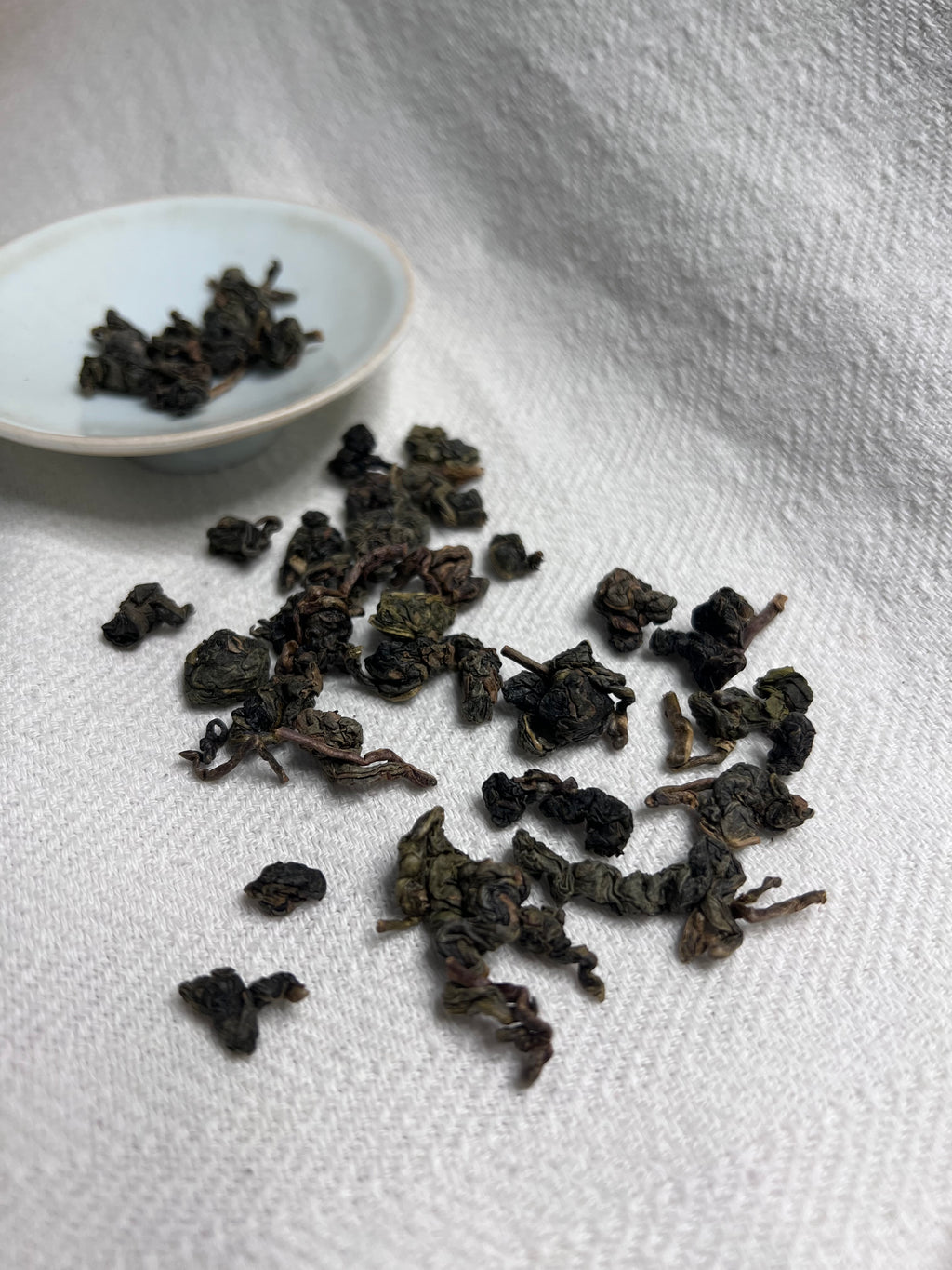 Rolled Da Ye oolong leaves scattered across white linen with a celadon saucer in the background