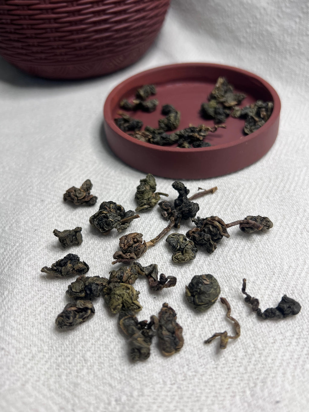 Tightly rolled Da Ye oolong leaves scattered on white linen beside a dark rose woven lacquer tea caddy